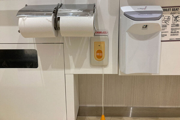 低い位置にある呼び出しボタンの写真、優先トイレ内で転倒した場合でも対応できるよう、低い位置からでも引っ張れるようにトイレットペーパーの右下に紐がついた呼び出しボタンがある
