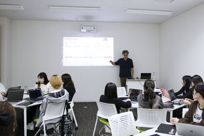 プロジェクターから投影された映像を背後に、大学生の受講者に向けて説明している尾野