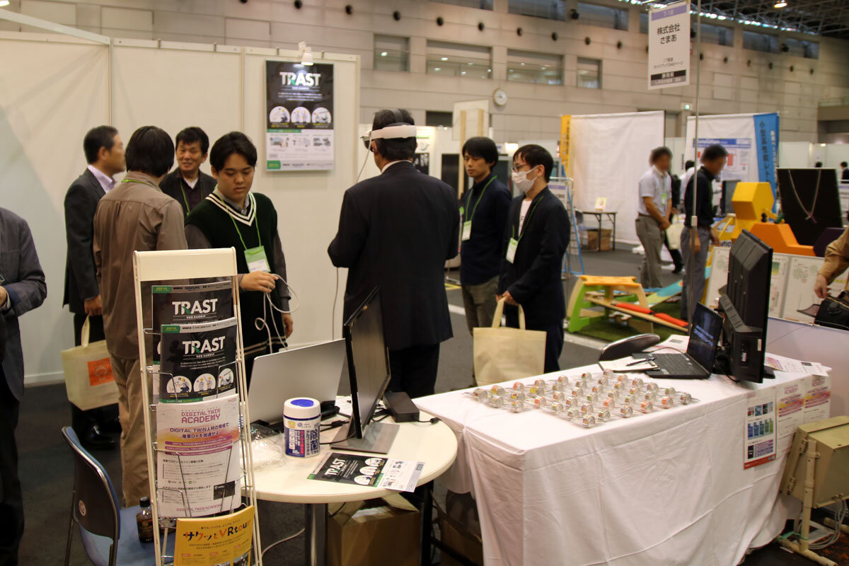 展示会の会場でのトラスト出展ブースの様子。NICOスタッフなど複数の男性が立っている中で、ひとり体験者の男性がヘッドマウントディスプレイを装着した状態で背中を向けて立っている