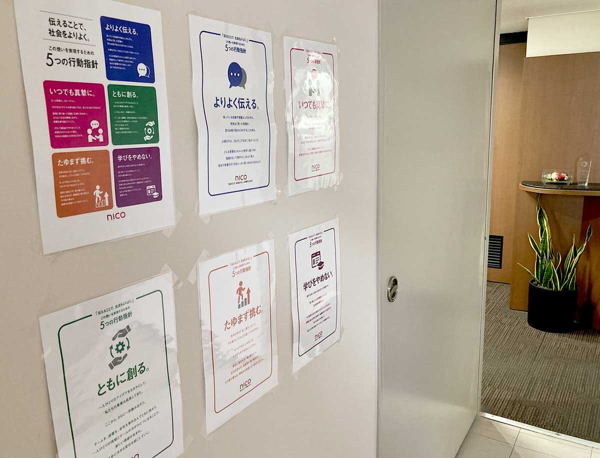 ビル内の壁に6枚のA4サイズのポスターが3列2行で掲出されている様子。左上の一枚は企業メッセージ、他5枚は行動指針が書かれている