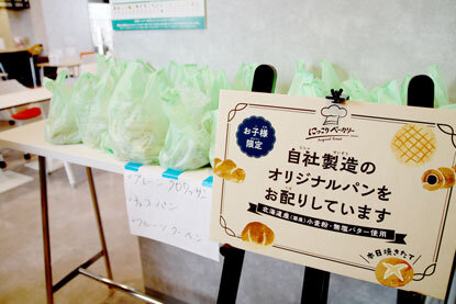 自社製造のオリジナルパンをお配りしていますという看板が立っており、その後ろにビニール袋に入ったパンがたくさん並んでいる