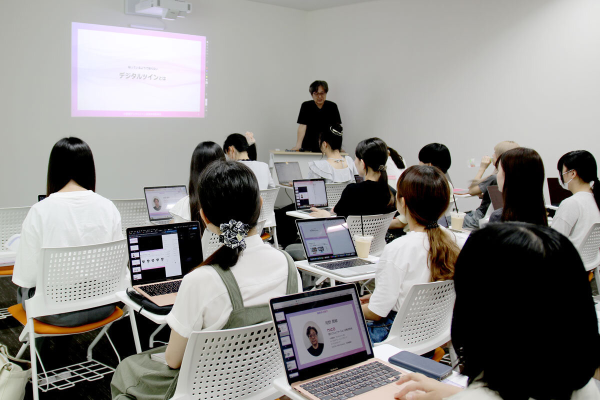 大学の講義室。椅子に座って講義を受ける大学生の後ろ姿と、それに向き合う形でスクリーンを背にしてしゃべる講師の姿