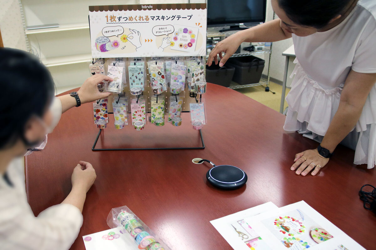 什器にかけられたbandeに手を伸ばすモニターと、それを見ているモデレーターの様子
。机の上にはむき出しのbandeも並んでいる