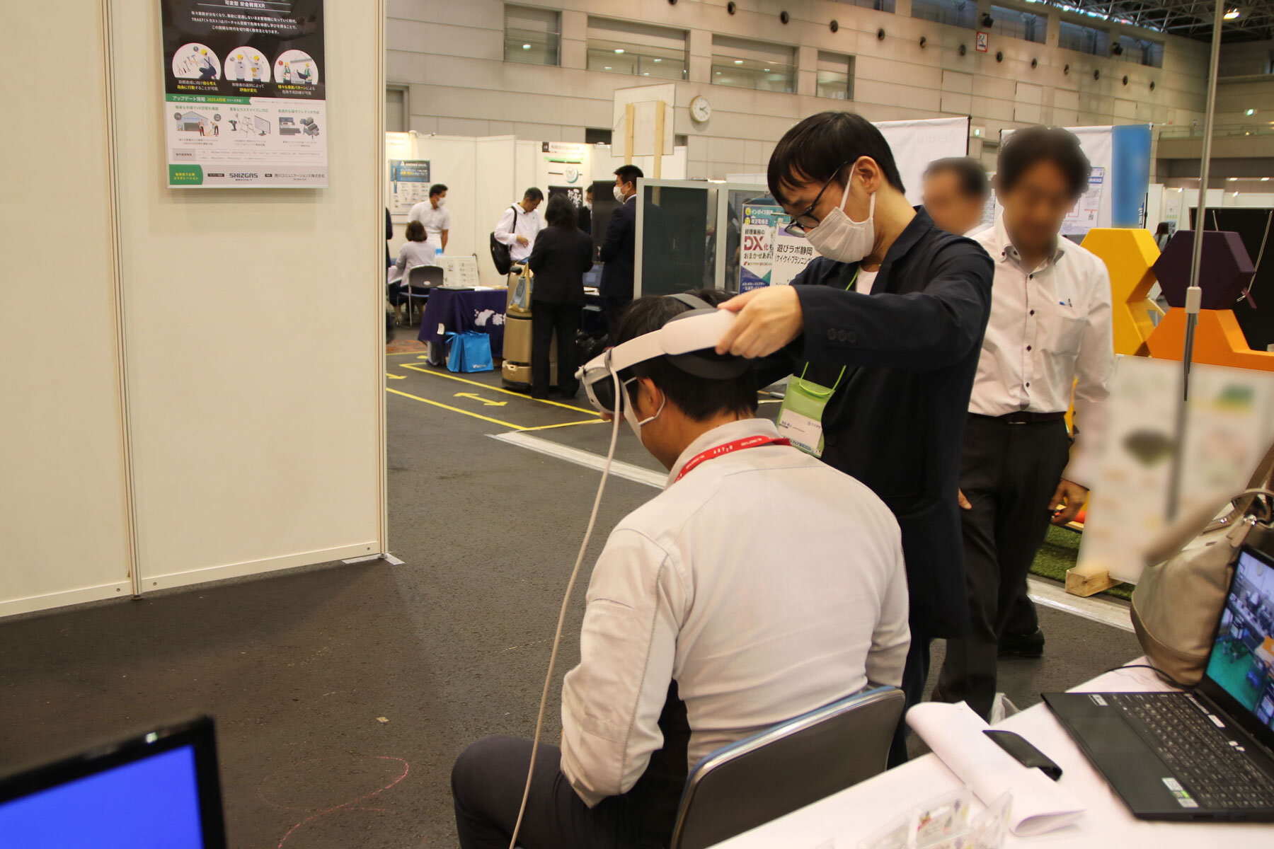 椅子に座って待つ体験者の男性と、その男性の頭にヘッドマウントディスプレイを装着しようとしている池谷の様子。
